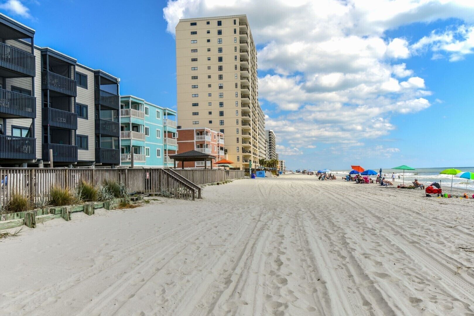 Een strand met een rij meerverdiepinggebouwen, waaronder een hoog beige gebouw en een lager blauw gebouw met balkons.