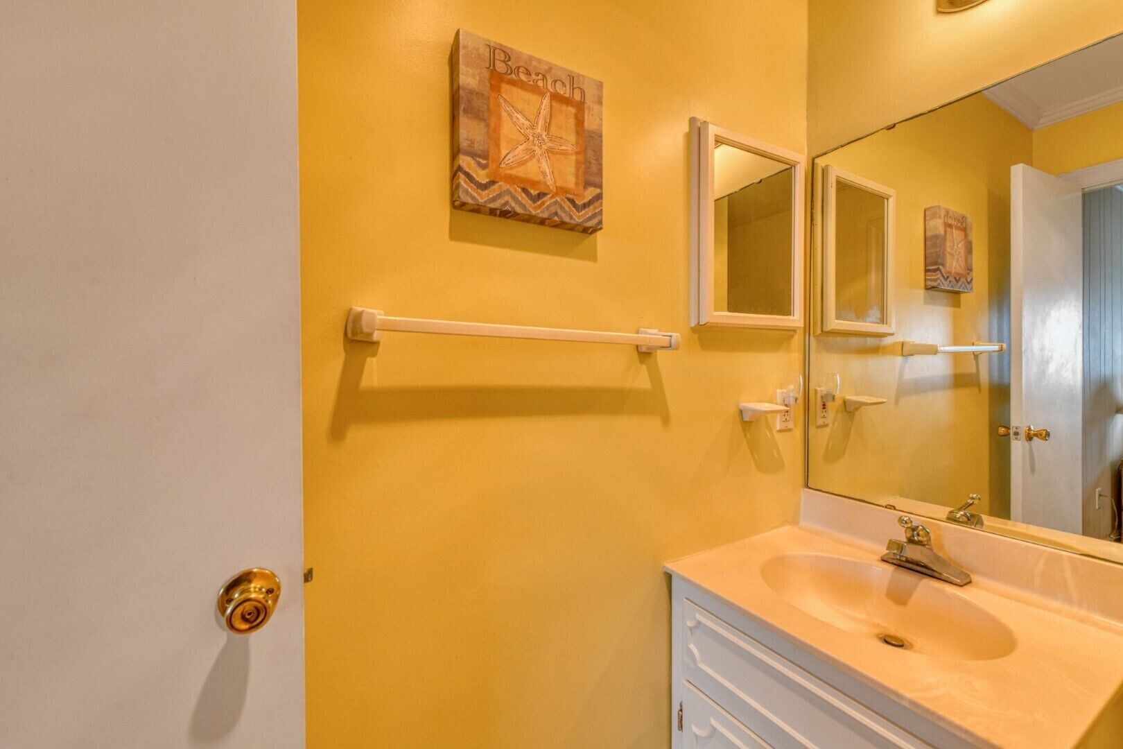 Ein Badezimmer mit gelber Wand, Waschbecken in Weiß, Spiegel und Handtuchhalter.