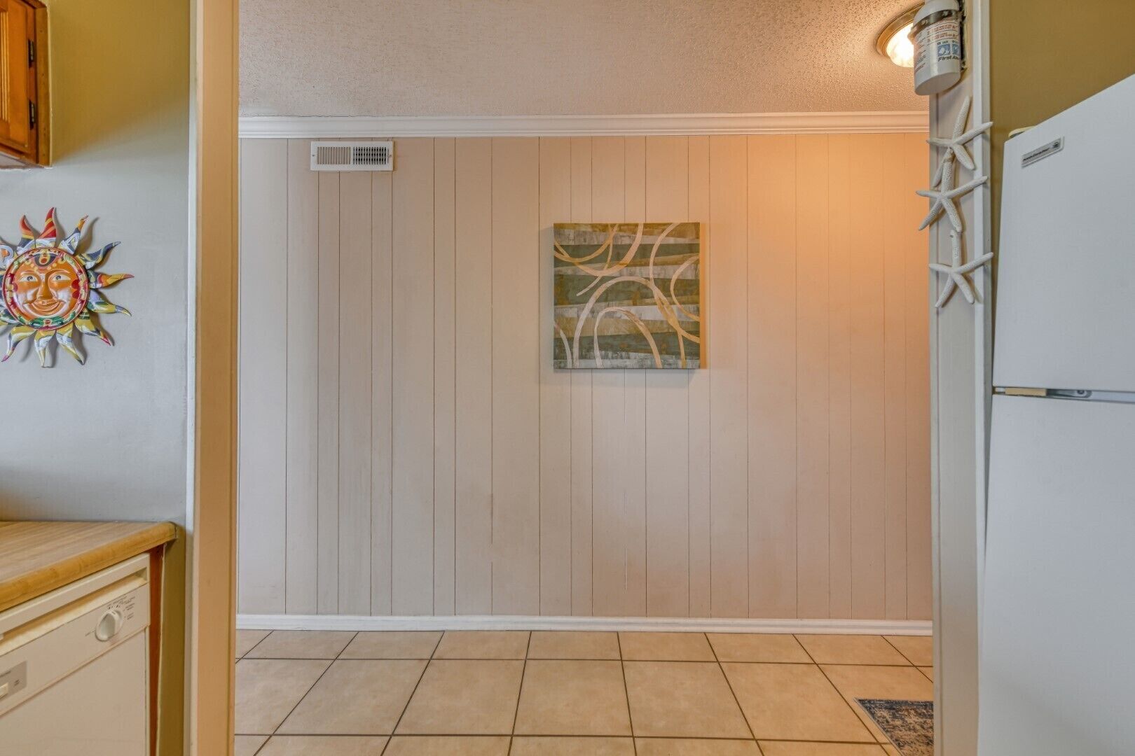 A kitchen with wooden panel walls, a tiled floor, a decorative sun wall art, and a ceiling vent.
