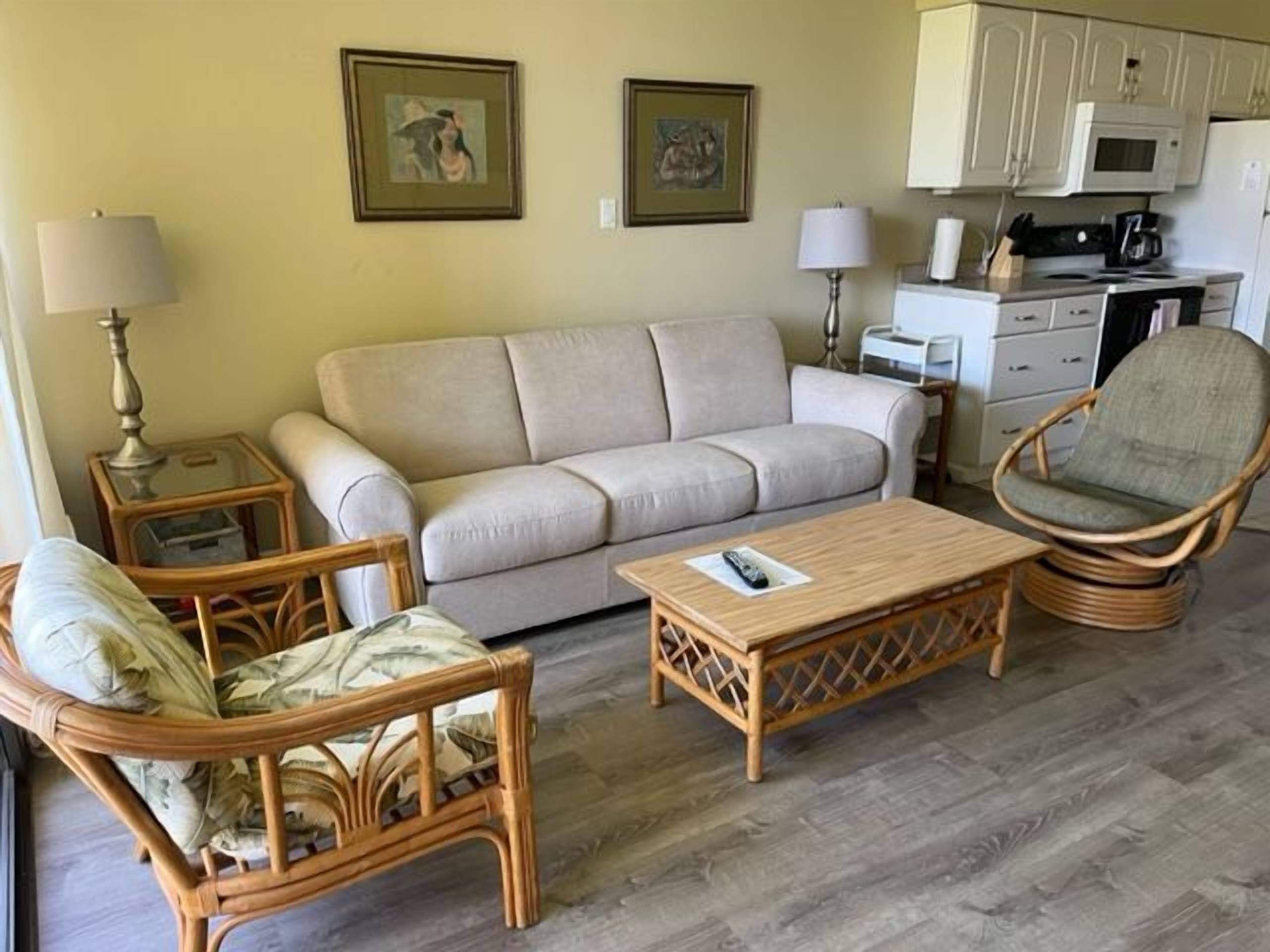 A living room with a sofa, a wicker chair, a wooden coffee table, and a kitchen area with a microwave and cabinets.
