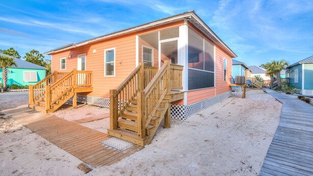 A beachfront house with a wooden staircase leading to a covered porch, a sandy deck, and a clear blue sky. - The Rookery III 7022 by RedAwning (Gulf Shores)