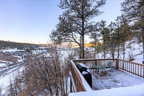 A deck with a table and chairs, overlooking a snowy landscape with trees and a river.