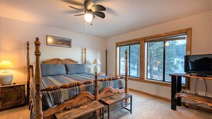 Un dormitorio con cama con dosel, ventilador de techo, televisor sobre un soporte de madera y un ventanal con vistas a árboles.
