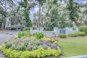 A landscaped garden with a variety of plants and flowers, a white building with a sign, and a street in the background.
