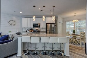 A modern kitchen with white cabinets, a central island with bar stools, stainless steel appliances, pendant lighting, and a clock on the wall.