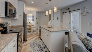 A modern kitchen with white cabinetry, granite countertops, and pendant lighting. There is a dining area with a table and chairs, and a built-in oven. - Sunset Rentals | Ocean Breeze 44 | Dog Friendly (Hilton Head Island)