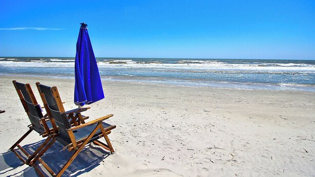 Ein Strand mit zwei Holzliegen und einem blauen Sonnenschirm.