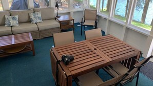 Een gezellig buitenzitgedeelte met een houten tafel en stoelen, een bank met gekleurde kussens en grote ramen met uitzicht op een meer.