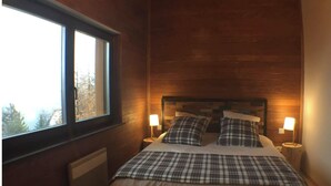 Een slaapkamer met een groot raam, houten wanden, een bed met geruit beddengoed, twee bedlampjes en een radiator.