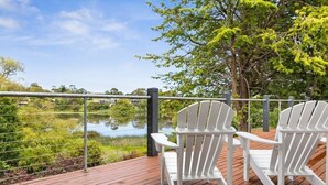 Two white Adirondack chairs on a wooden deck overlooking a body of water with a metal railing and trees in the background. - Bellarine Lake House Ocean Grove (Ocean Grove)