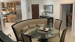 Un comedor moderno con una mesa de vidrio rodeada de sillas, una cocina al fondo y una puerta que da a otra habitación.