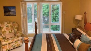 Un dormitorio con un ventanal grande, una cama con colcha a rayas, una silla con estampado de piña y un cuadro en la pared.