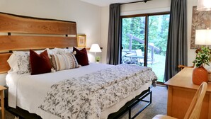 A bedroom with a large bed, a wooden headboard, a nightstand, a desk with a chair, a potted plant, and a view of a deck and trees through a large window. - Windfall @ Quechee Club VT: Cozy Updated Condo  A/C, Fireplace -Ski -Golf -Renew (Hartford)