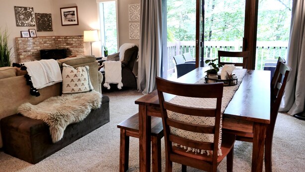 A cozy living room with a fireplace, a dining table with chairs, and a view of a balcony with a wooden railing. - Windfall @ Quechee Club VT: Cozy Updated Condo  A/C, Fireplace -Ski -Golf -Renew (Hartford)