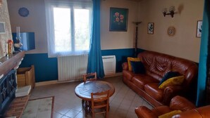 A living room with a brown leather sofa, a dining table with chairs, a window with blue curtains, and a radiator.