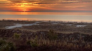 Un paisaje costero con un atardecer sobre el océano, terreno arenoso y vegetación escasa.