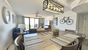 Un comedor con una mesa de madera, sillas y individuales tejidos. Hay un ventilador de techo, una bicicleta y un reloj en la pared.