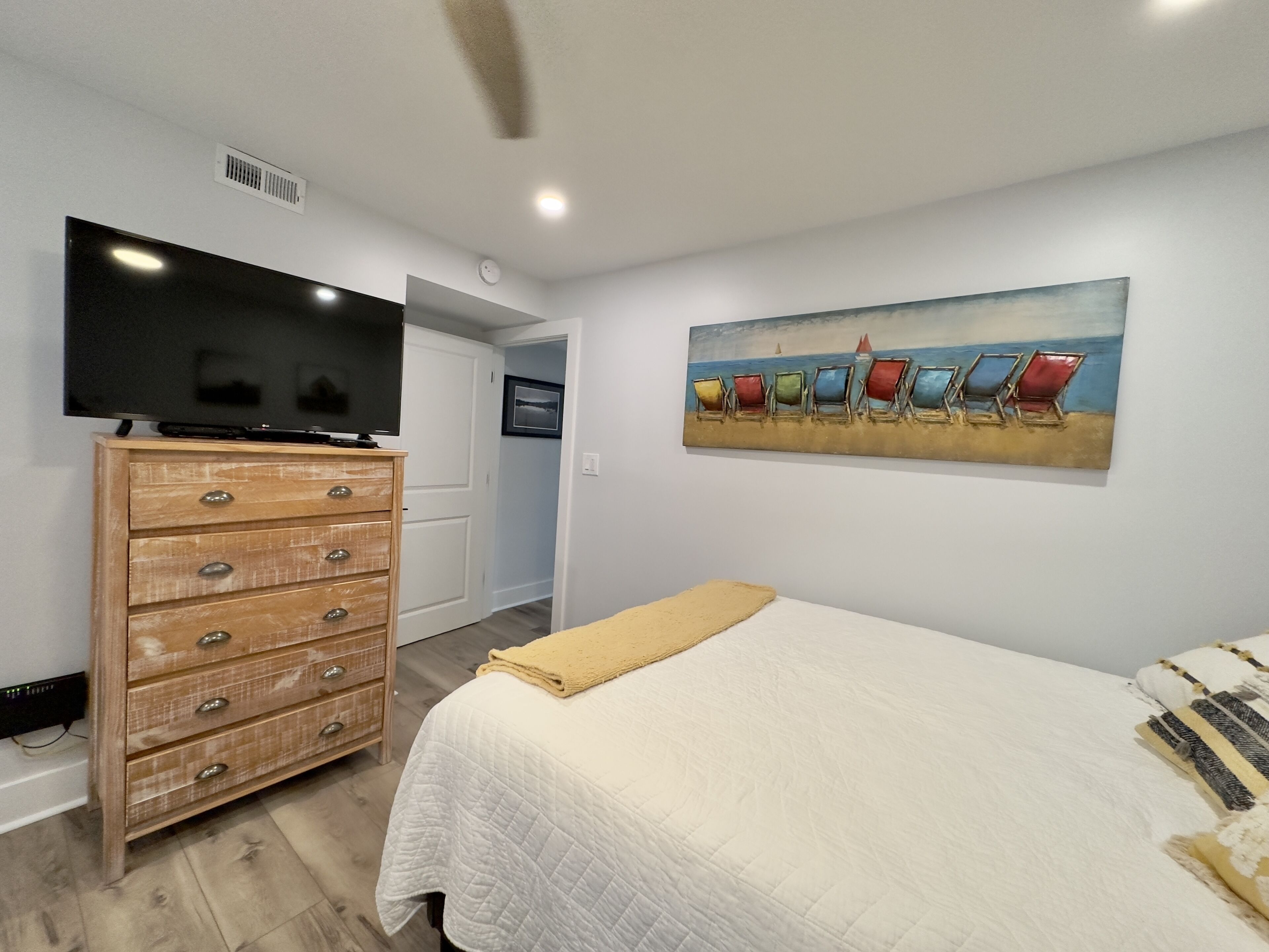 A bedroom with a bed, a dresser, a television, and a painting of beach chairs.