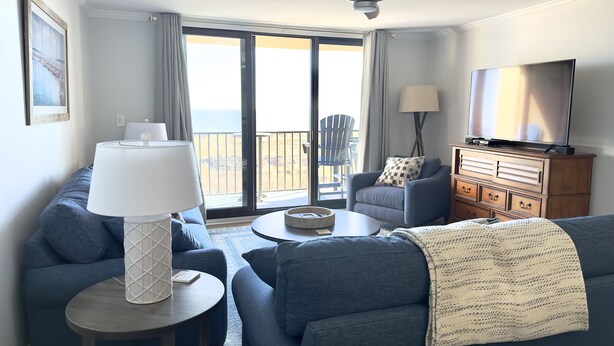 Una sala moderna con sofá, sillón y televisor. Hay un balcón con vista al océano.