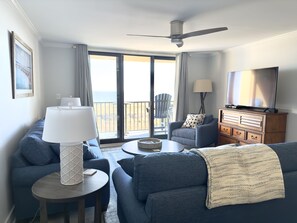 Een moderne woonkamer met een bank, fauteuil en een televisie. Er is een balkon met uitzicht op de oceaan.