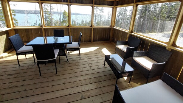 A wooden deck with a glass enclosure, featuring a table and chairs, overlooking a body of water and forested area. - House with Stunning Ocean View (Mulgrave)