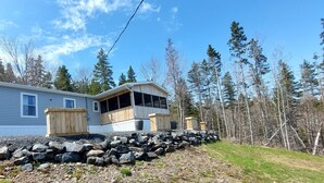 A cabin with a large deck, a stone retaining wall, and a forested backdrop. - House with Stunning Ocean View (Mulgrave)
