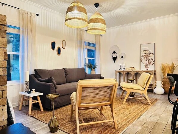 A cozy living room with a brown sofa, a wooden chair, and a small side table. There are two hanging lamps, a vase with dried plants, and a framed picture on the wall.