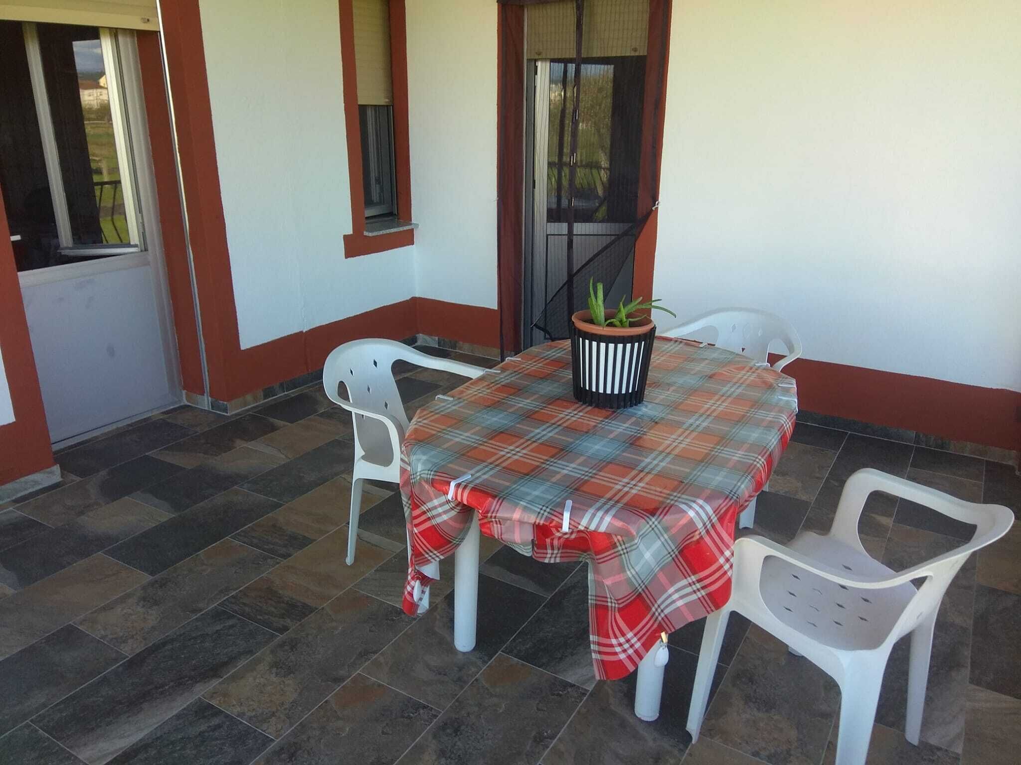 A small outdoor patio with a table covered in a plaid tablecloth, surrounded by white plastic chairs.