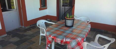 A small outdoor patio with a table covered in a plaid tablecloth, surrounded by white plastic chairs.