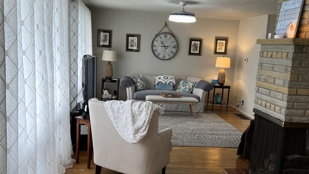 Una sala de estar con sofá, sillón y chimenea. Hay un reloj en la pared y una lámpara sobre una mesita.