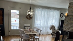 Un comedor con una mesa redonda y sillas, una araña y vistas a través de una ventana con cortinas.