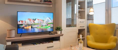 A living room with a TV displaying a harbor scene, wooden furniture, a yellow armchair, and a glass cabinet.