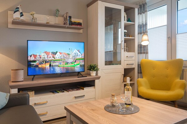 A living room with a TV displaying a harbor scene, wooden furniture, a yellow armchair, and a glass cabinet.