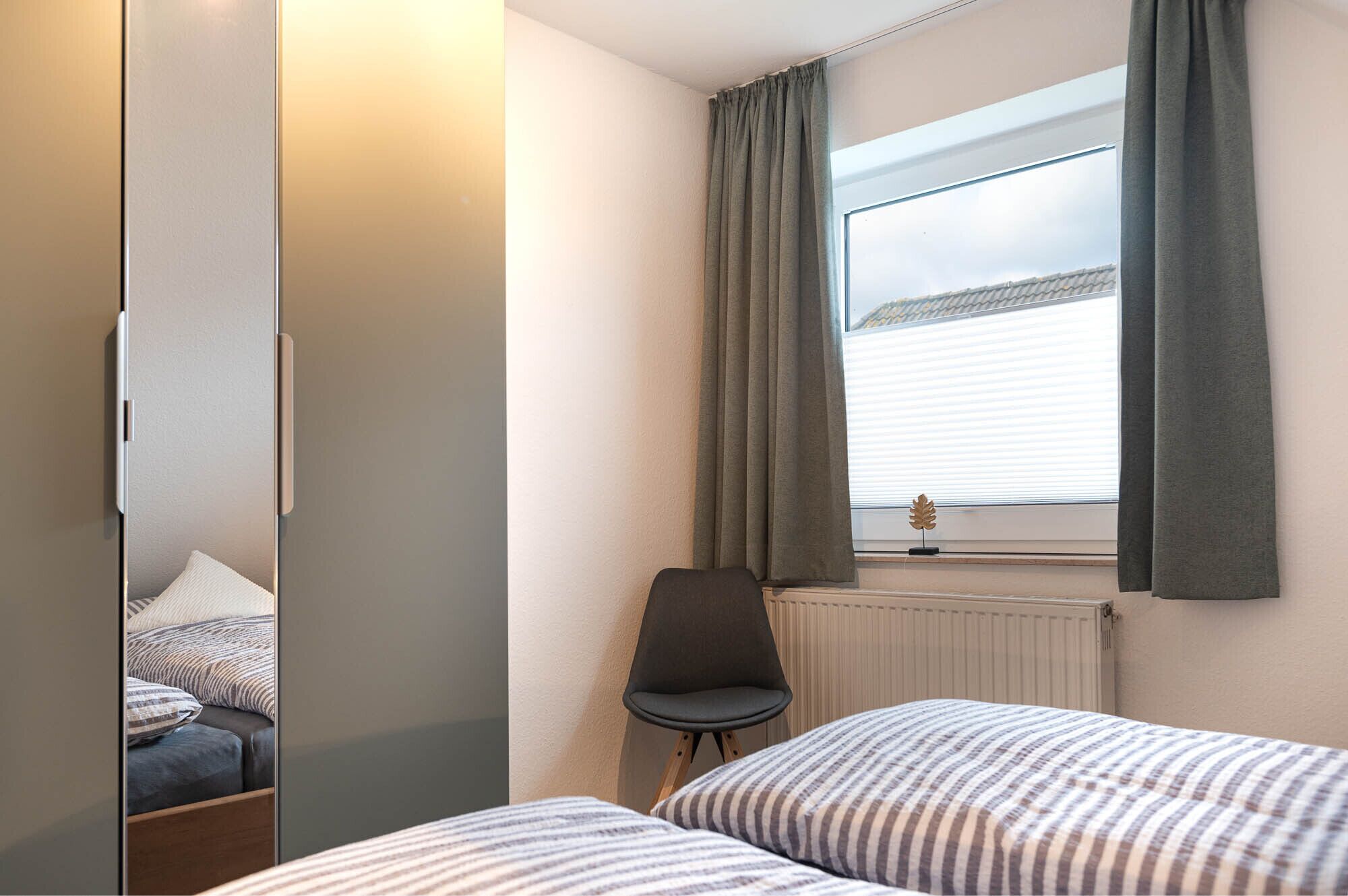 A bedroom with two beds, a chair, a radiator, and a window with curtains.
