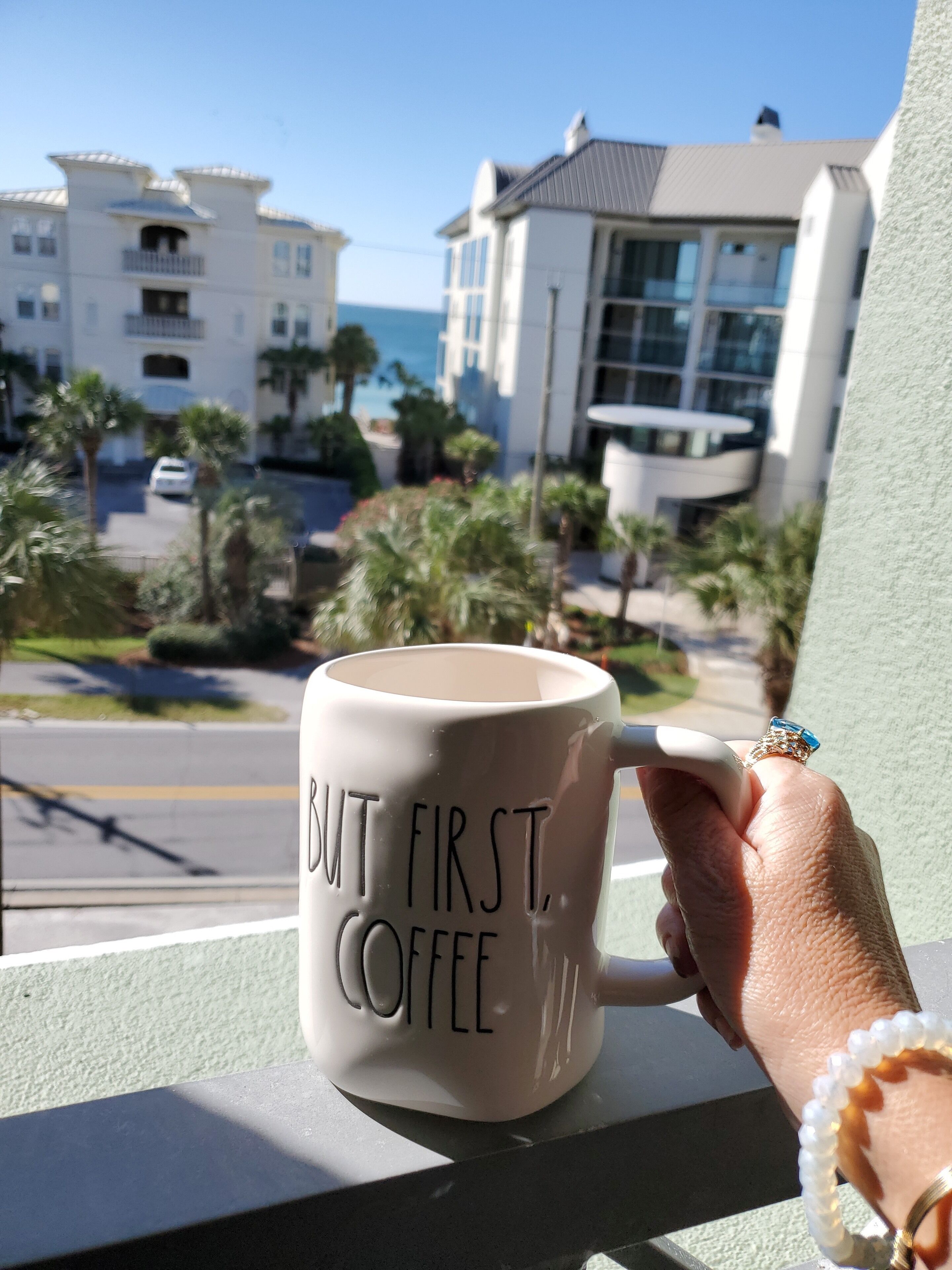 A hand holding a mug with the text "BUT FIRST, COFFEE" on a balcony overlooking a residential area with palm trees and buildings.