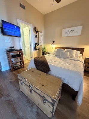 A bedroom with a bed, a wall-mounted TV, a guitar, a plant, and a suitcase.