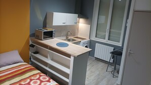 Een compacte kamer met een kitchenette, een bed met een bed met een gestreept deken en een raam met zonwering.