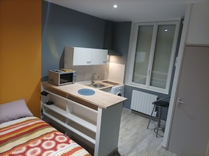 Quarto compacto com cozinha compacta, cama com manta estampada e janela com persianas.