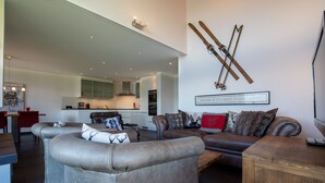 A modern living room with a brown leather sofa, a grey armchair, and a wooden coffee table. There is a kitchen in the background with white cabinets and a dining area with a red chair. - Ski Paradise SP 011 - MOUNTAIN apartment 6 pers (Veysonnaz)