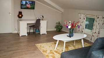 Ett vardagsrum med ett vitt skrivbord, en tv som är monterad på väggen, en vas med blommor på ett bord och ett fönster med gardiner.