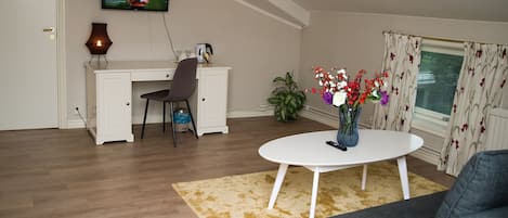Un salón con un escritorio blanco, un televisor montado en la pared, un jarrón con flores sobre una mesa y una ventana con cortinas.