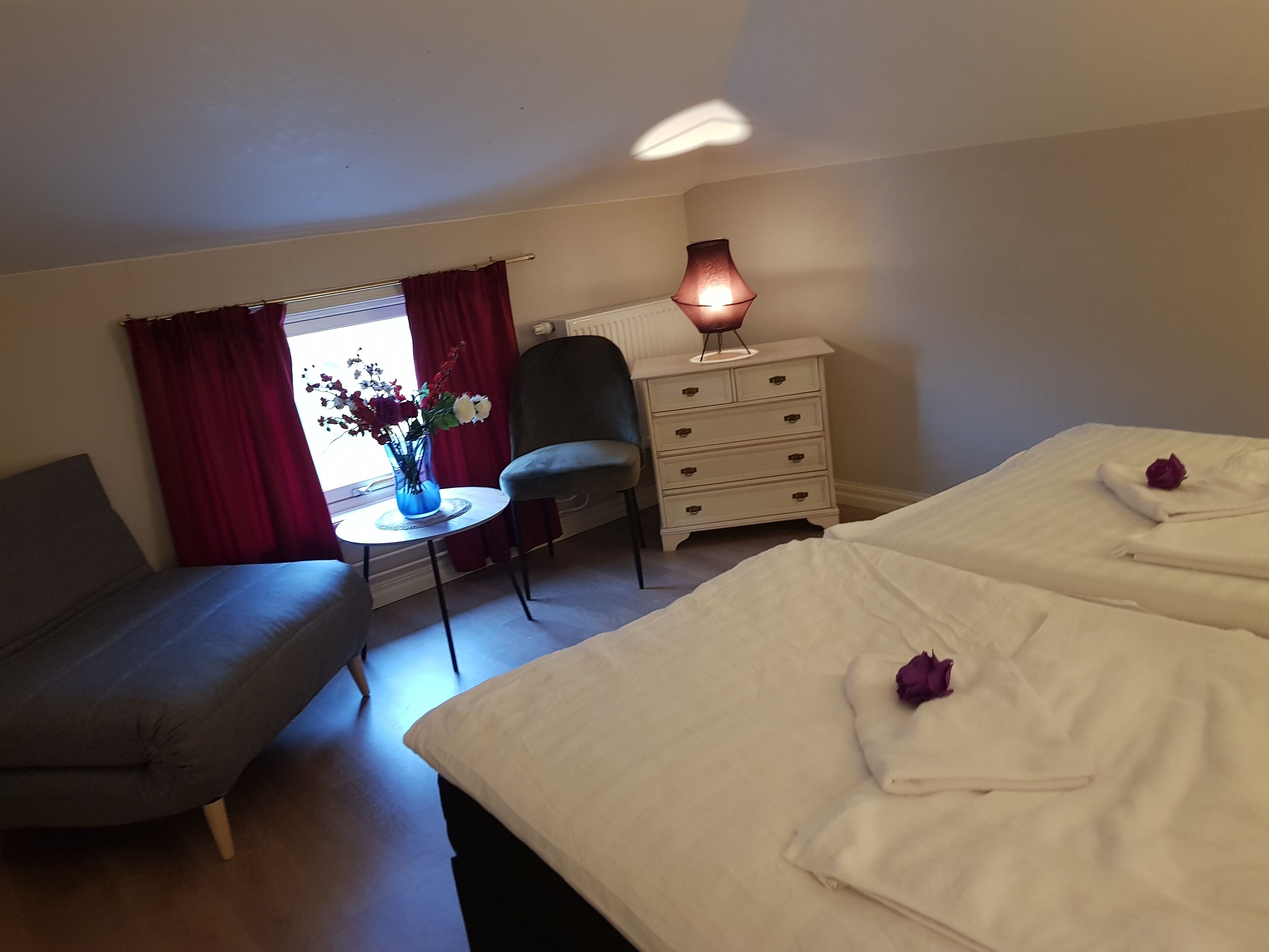 Habitación de hotel con dos camas, una silla, una mesita con un florero y un aparador.