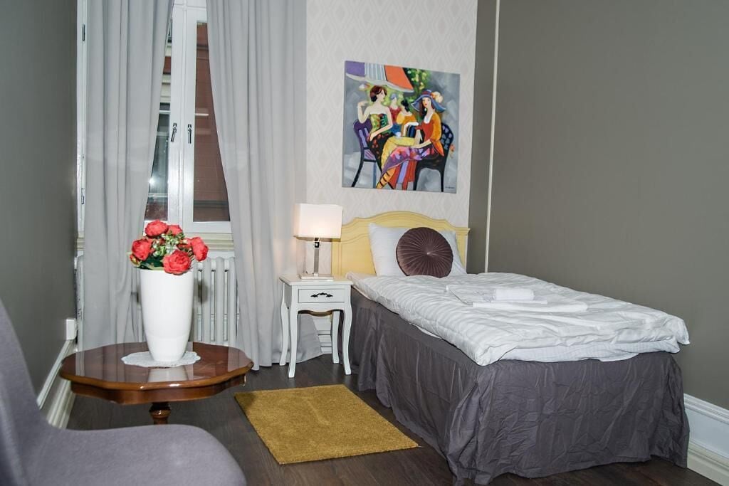 Una habitación de hotel con una cama, una mesita de noche con una lámpara, una pequeña mesa con un florero y un cuadro en la pared.