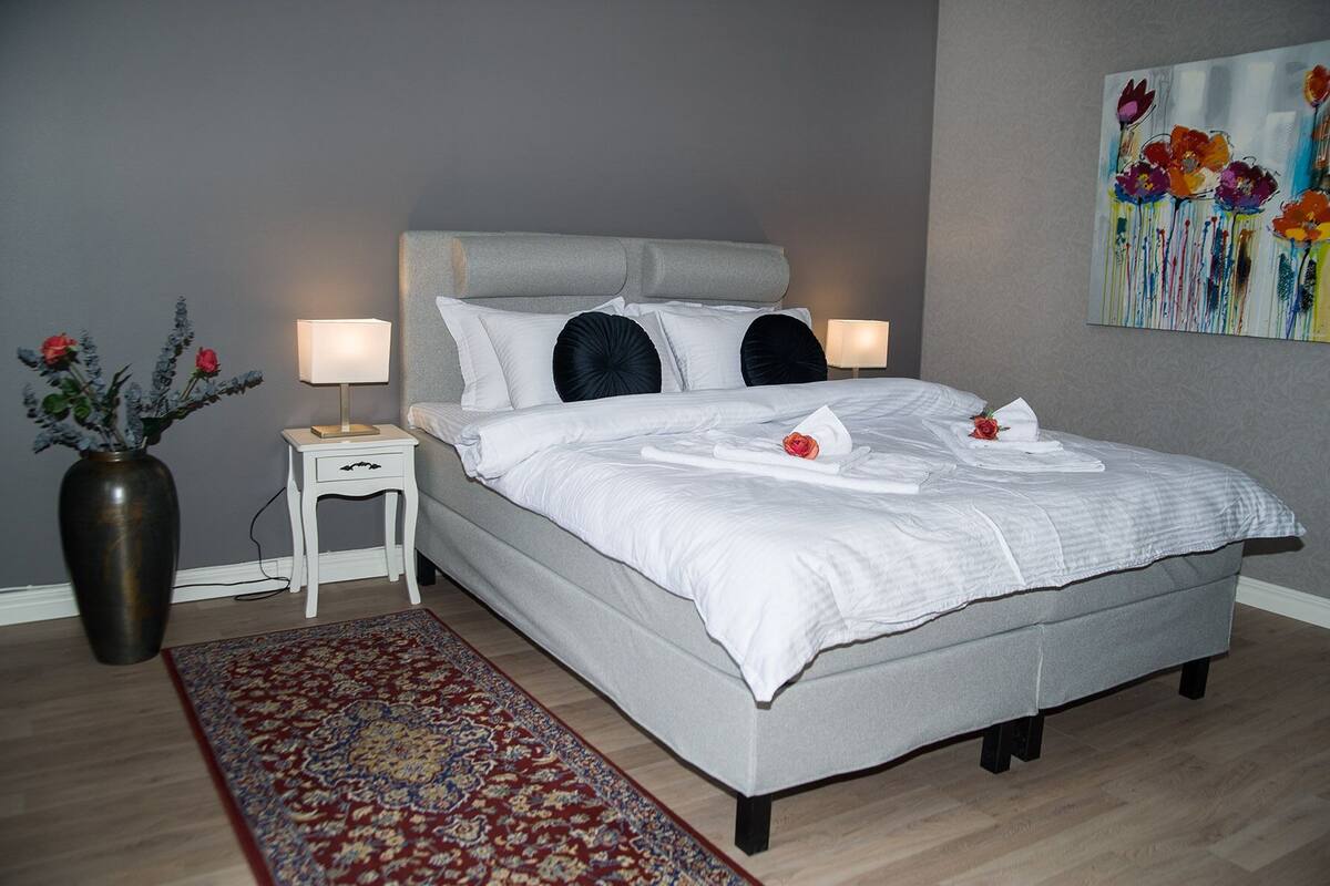 A neatly made bed with white linens and two pillows, a bedside table with a lamp, a vase with flowers, and a painting on the wall.