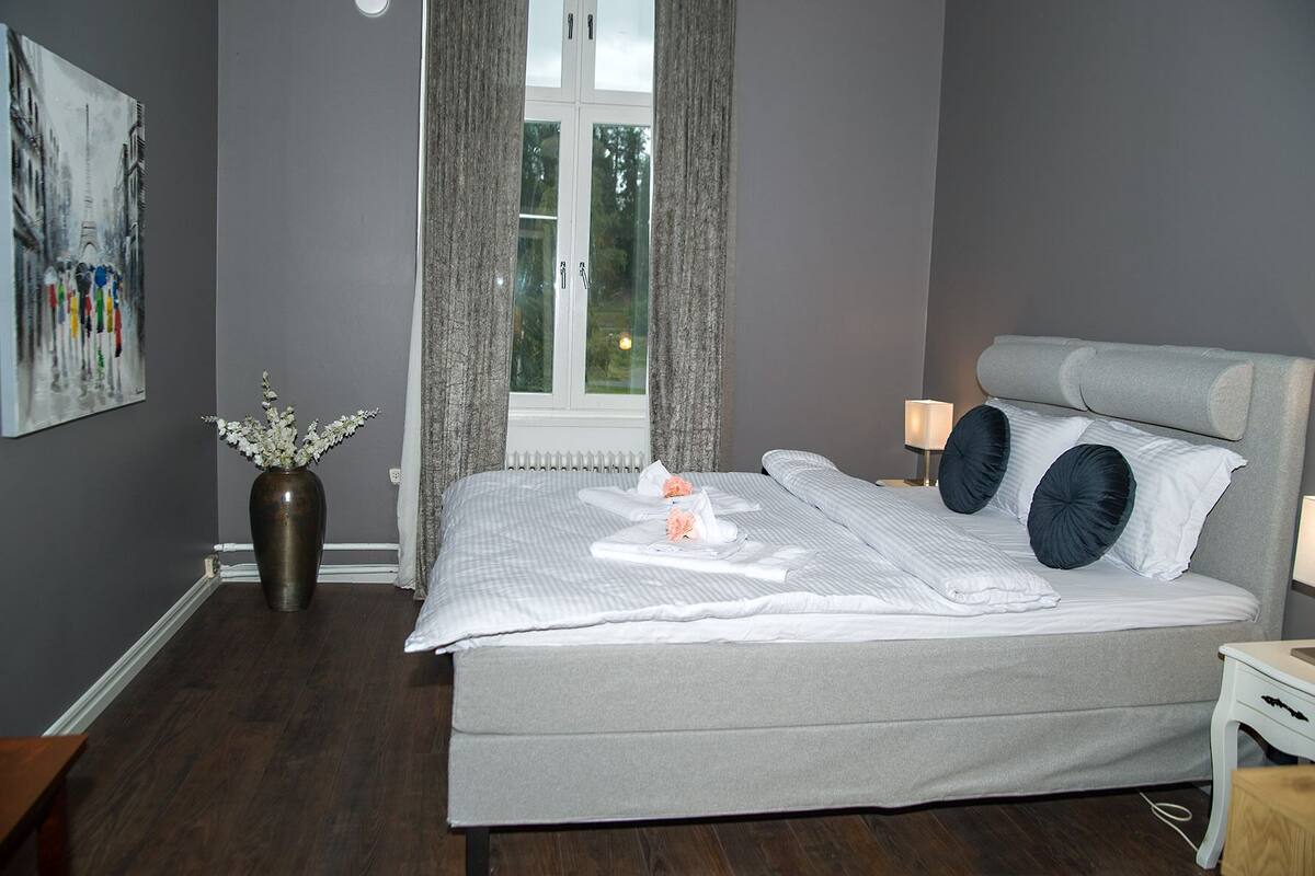 A modern bedroom with a large bed, a vase with flowers, a painting on the wall, and a window with curtains.