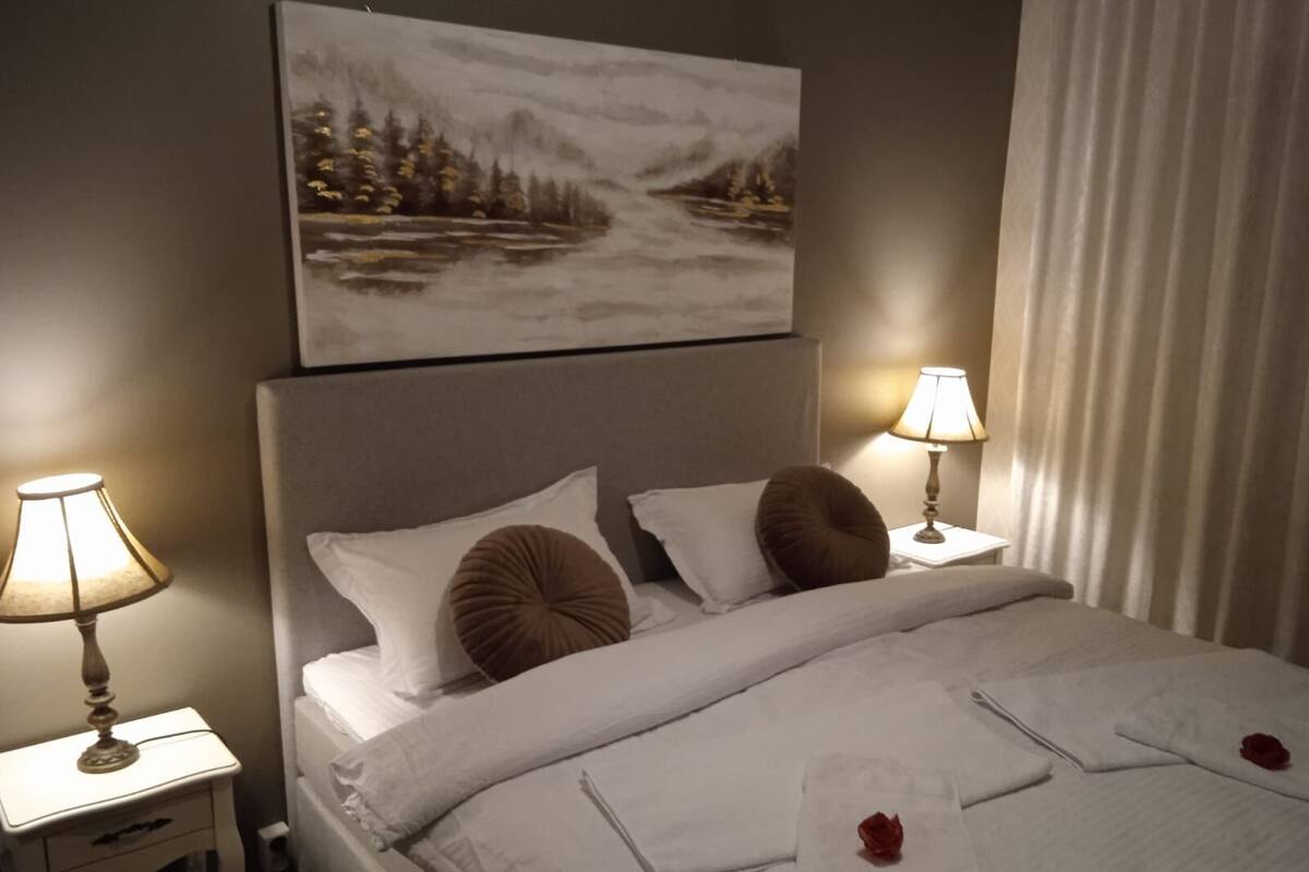 A hotel room with a bed, two bedside lamps, a painting above the bed, and a window with curtains.