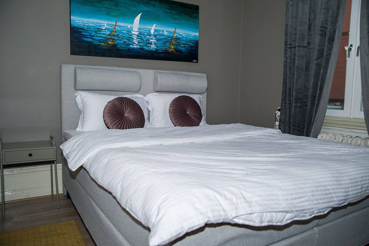 A neatly made bed with a headboard, a small bedside table, and a painting of sailboats on the wall.