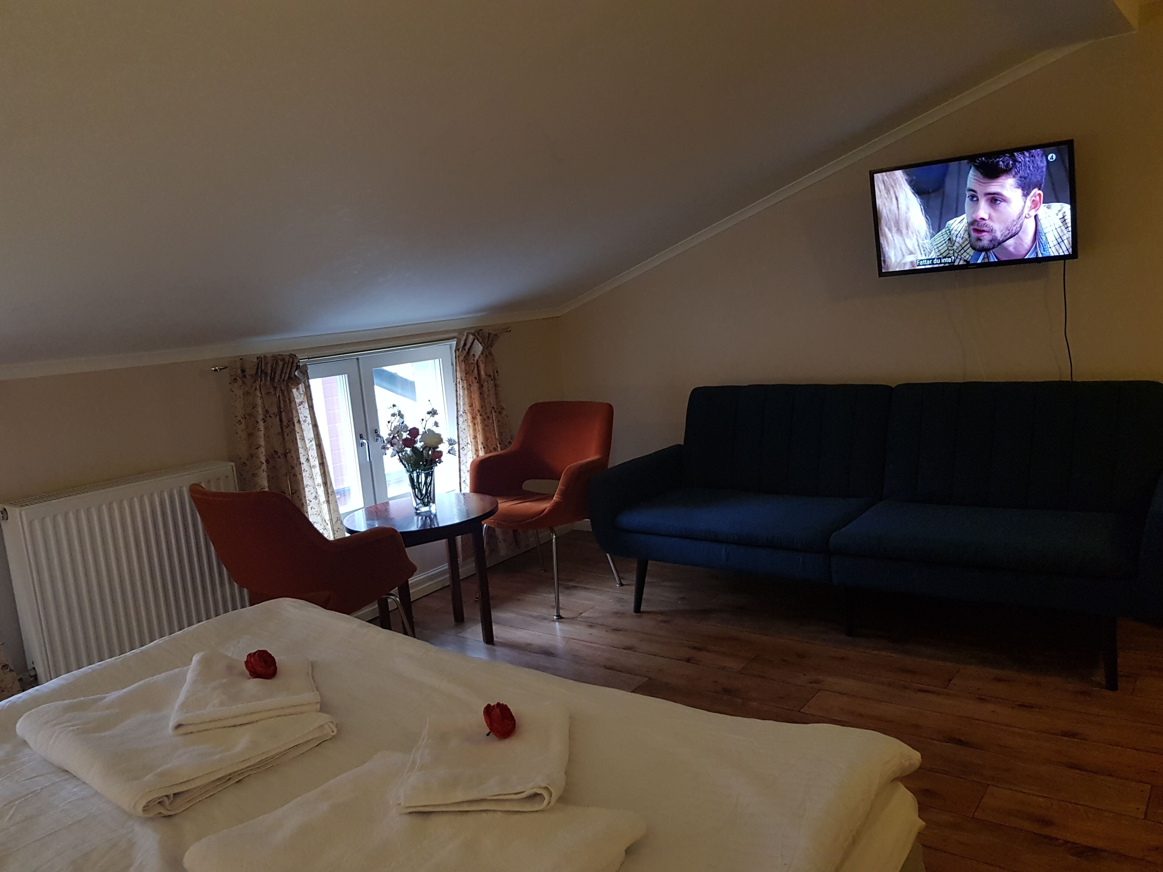 A room with a dark blue sofa, a red armchair, a small table with a vase of flowers, and a bed with folded white towels and a red rose. A television is mounted on the wall.