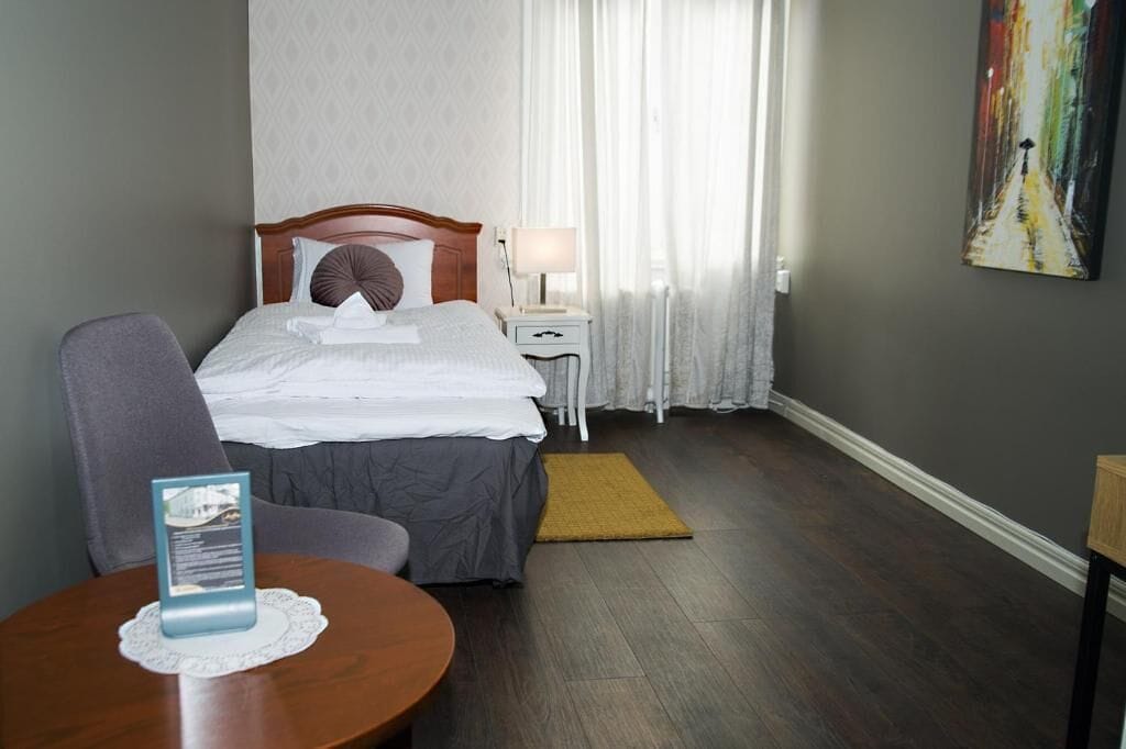 A hotel room with a bed, a chair, a small table, a nightstand, and a painting on the wall.