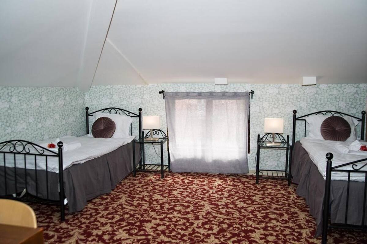 A room with two beds, each with a nightstand and a lamp. The room has patterned wallpaper and a red carpet.